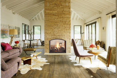 Imagen de salón campestre con paredes blancas, suelo de madera en tonos medios, chimenea de doble cara y marco de chimenea de piedra