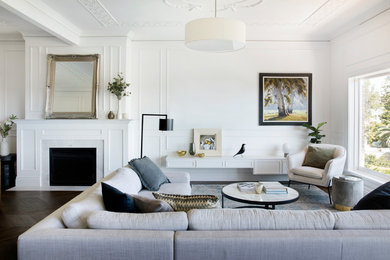 На фото: гостиная комната в стиле неоклассика (современная классика) с белыми стенами, темным паркетным полом, стандартным камином и коричневым полом с
