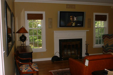 Modelo de salón clásico de tamaño medio con paredes amarillas, suelo de madera en tonos medios, todas las chimeneas, marco de chimenea de madera y televisor colgado en la pared