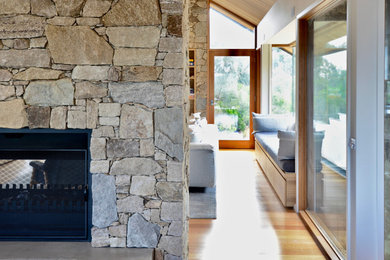 Cette image montre un salon marin de taille moyenne et ouvert avec un mur blanc, un sol en bois brun, une cheminée double-face, un manteau de cheminée en pierre, un téléviseur fixé au mur et un sol marron.