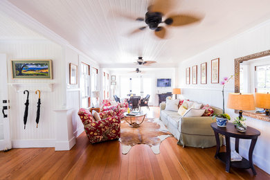 Imagen de salón abierto tropical de tamaño medio con paredes blancas, suelo de madera en tonos medios y televisor colgado en la pared