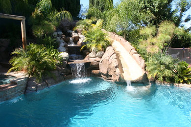 Modelo de piscina con tobogán alargada tropical de tamaño medio a medida en patio trasero con adoquines de piedra natural