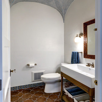 Powder room - mid-sized mediterranean terra-cotta tile and multicolored floor powder room idea in Los Angeles with open cabinets, a trough sink, medium tone wood cabinets, a two-piece toilet, white walls and solid surface countertops