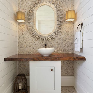 Cette image montre un WC et toilettes ethnique avec un placard à porte plane, des portes de placard blanches, un carrelage multicolore, une plaque de galets, un mur blanc, un sol en galet, une vasque, un plan de toilette en bois, un sol multicolore et un plan de toilette marron.