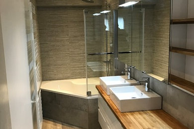 Cette image montre une petite salle de bain principale design avec une baignoire d'angle, un carrelage gris, un mur gris, un lavabo posé, un plan de toilette en bois et un sol marron.