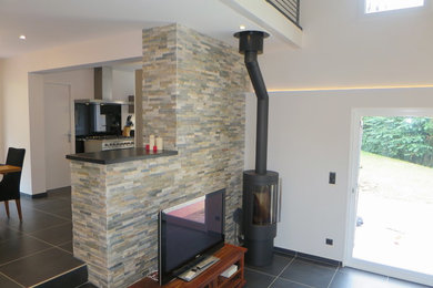 Large trendy loft-style ceramic tile and black floor family room photo in Lyon with gray walls, a wood stove, a stone fireplace and a tv stand