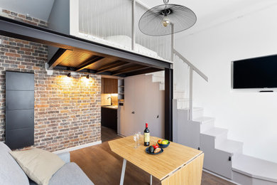 Modelo de sala de estar abierta urbana de tamaño medio sin chimenea con paredes blancas, suelo de madera oscura, televisor colgado en la pared, suelo marrón y ladrillo