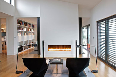 Modelo de biblioteca en casa abierta contemporánea grande sin televisor con paredes blancas, suelo de madera clara, chimenea lineal y marco de chimenea de metal