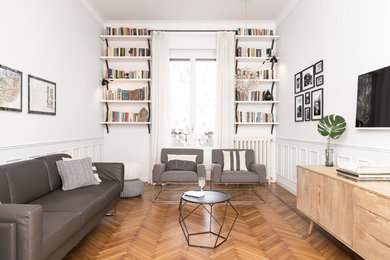 Modelo de biblioteca en casa gris y blanca escandinava de tamaño medio con televisor colgado en la pared, paredes blancas, suelo de madera en tonos medios y suelo marrón