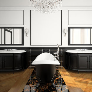 Aménagement d'une très grand salle de bain principale classique avec un placard avec porte à panneau encastré, des portes de placard noires, une baignoire sur pieds, un sol en bois brun, un lavabo intégré et un mur blanc.