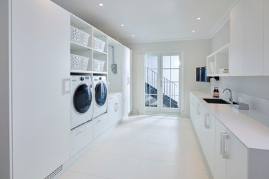 Multifunktionaler, Zweizeiliger Moderner Hauswirtschaftsraum mit Waschbecken, weißer Wandfarbe, Keramikboden, Waschmaschine und Trockner nebeneinander, flächenbündigen Schrankfronten, weißen Schränken und Mineralwerkstoff-Arbeitsplatte in London