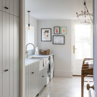 This is an example of a large classic single-wall utility room in London with a belfast sink, louvered cabinets, grey cabinets, white walls, porcelain flooring, a side by side washer and dryer, grey floors and white worktops.