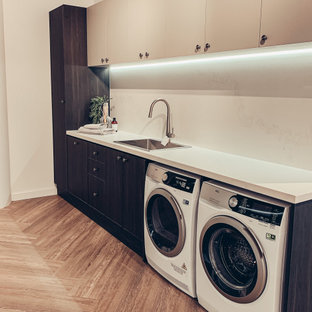 Esempio di una sala lavanderia design di medie dimensioni con lavello a vasca singola, ante con riquadro incassato, ante in legno bruno, top in quarzo composito, paraspruzzi bianco, paraspruzzi in quarzo composito, pareti bianche, pavimento in laminato, lavatrice e asciugatrice affiancate, pavimento marrone, top bianco e pannellatura