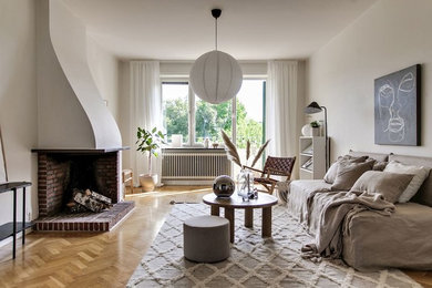Imagen de salón nórdico con paredes blancas, suelo de madera en tonos medios, chimenea de esquina, marco de chimenea de ladrillo y suelo beige