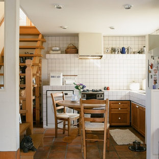 Kleine Country Wohnküche ohne Insel in L-Form mit profilierten Schrankfronten, hellbraunen Holzschränken, Arbeitsplatte aus Fliesen, weißen Elektrogeräten und Backsteinboden in Sonstige