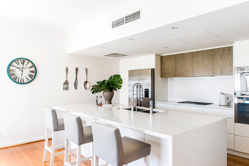 Photo of a beach style galley kitchen in Brisbane with a double-bowl sink, flat-panel cabinets, white cabinets, white splashback, mosaic tile splashback, stainless steel appliances, medium hardwood floors and with island.