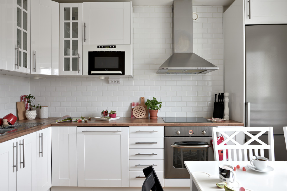 Photo of a scandinavian l-shaped eat-in kitchen in Other with shaker cabinets, white cabinets, wood benchtops, white splashback, subway tile splashback, stainless steel appliances and brown benchtop.