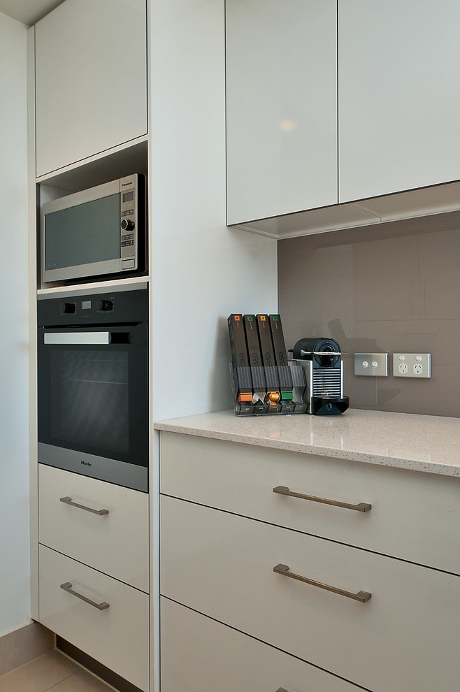 This is an example of a small modern l-shaped separate kitchen in Gold Coast - Tweed with an undermount sink, flat-panel cabinets, white cabinets, quartz benchtops, grey splashback, glass sheet splashback, stainless steel appliances, ceramic floors and no island.