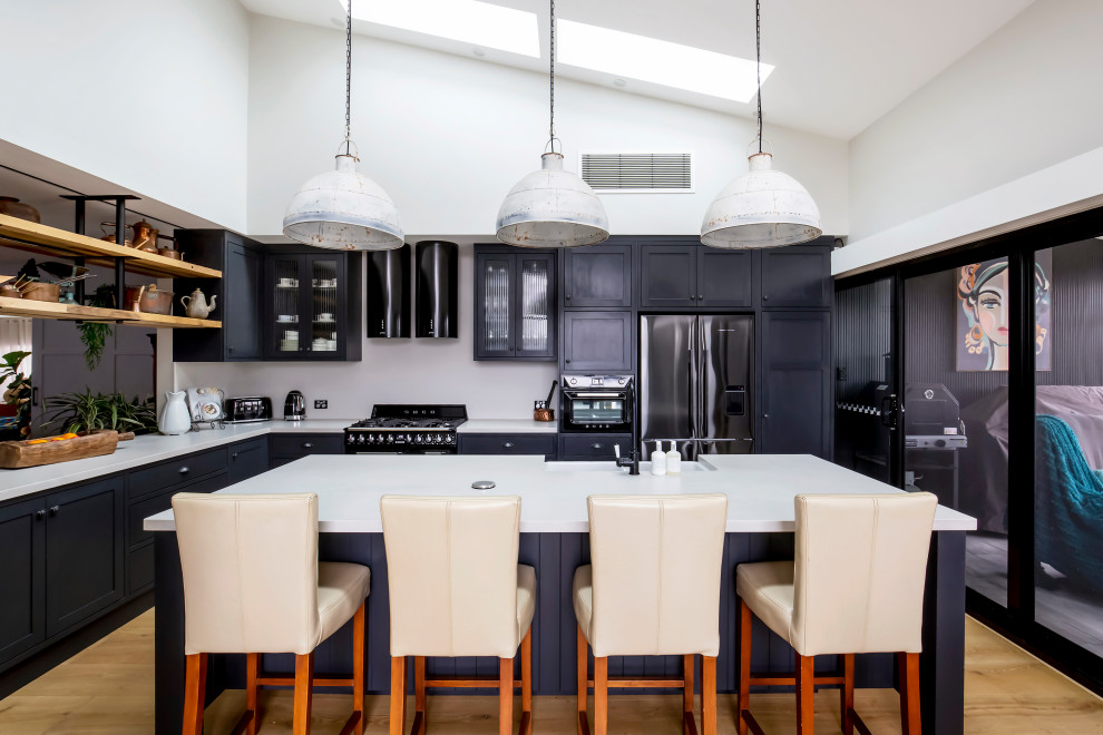 Photo of a large transitional l-shaped open plan kitchen in Sydney with a farmhouse sink, shaker cabinets, black cabinets, quartz benchtops, white splashback, engineered quartz splashback, black appliances, light hardwood floors, with island, white benchtop and vaulted.