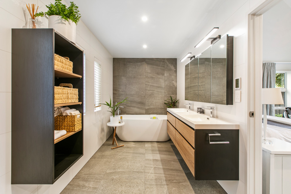 Photo of a contemporary master bathroom in Auckland with flat-panel cabinets, medium wood cabinets, a freestanding tub, gray tile, white walls, an integrated sink, grey floor and white benchtops.