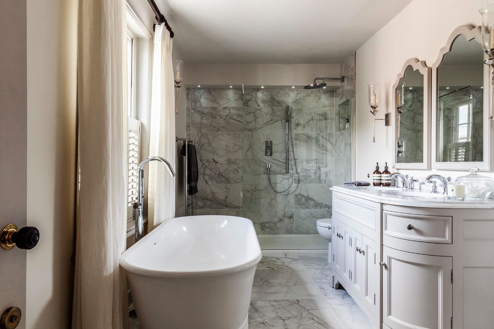 Photo of a mid-sized country master bathroom in Surrey with grey cabinets, a freestanding tub, a one-piece toilet, gray tile, marble, marble floors, an undermount sink, marble benchtops, grey floor, white benchtops, recessed-panel cabinets, a curbless shower, beige walls and a hinged shower door.
