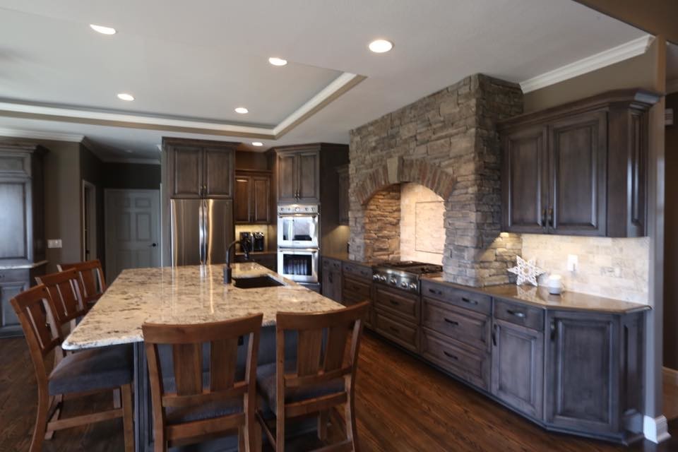 This is an example of a large country u-shaped eat-in kitchen in Kansas City with a single-bowl sink, raised-panel cabinets, grey cabinets, granite benchtops, brown splashback, travertine splashback, stainless steel appliances, dark hardwood floors, with island, brown floor and brown benchtop.