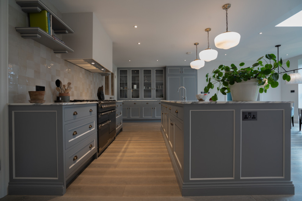 Design ideas for a medium sized contemporary kitchen/diner in London with a belfast sink, shaker cabinets, grey cabinets, stone tiled splashback, integrated appliances, light hardwood flooring, an island, brown floors, white worktops and feature lighting.