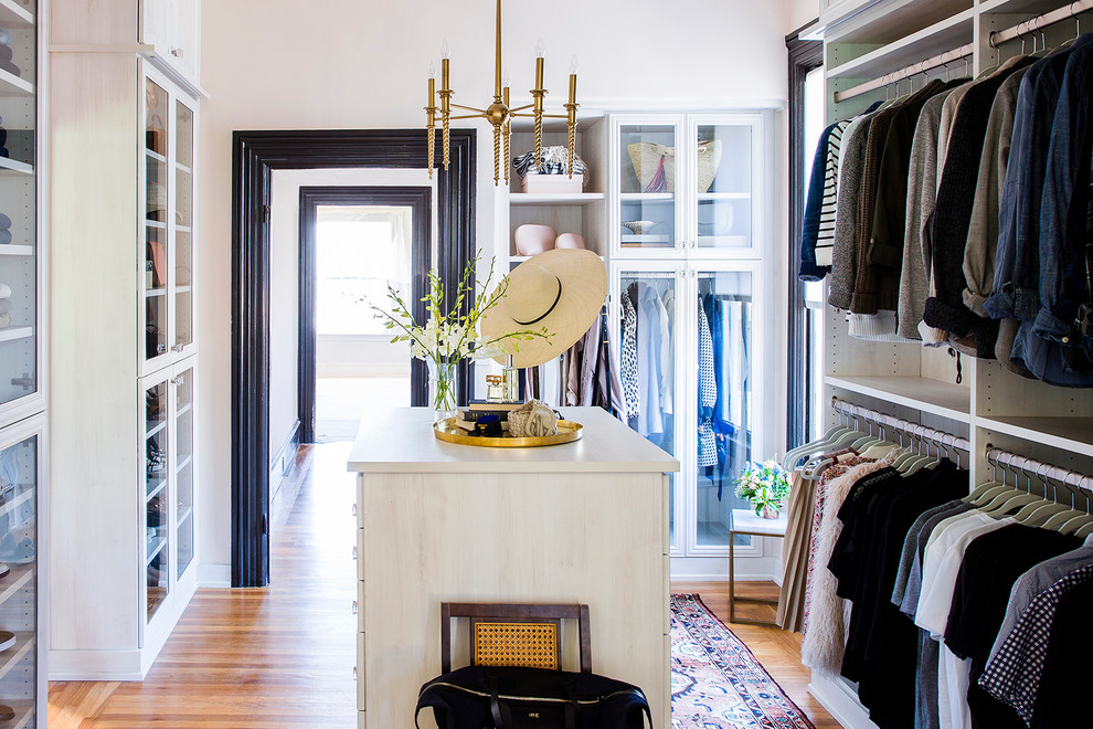 This is an example of an expansive transitional gender-neutral walk-in wardrobe in Other with glass-front cabinets, white cabinets and light hardwood floors.