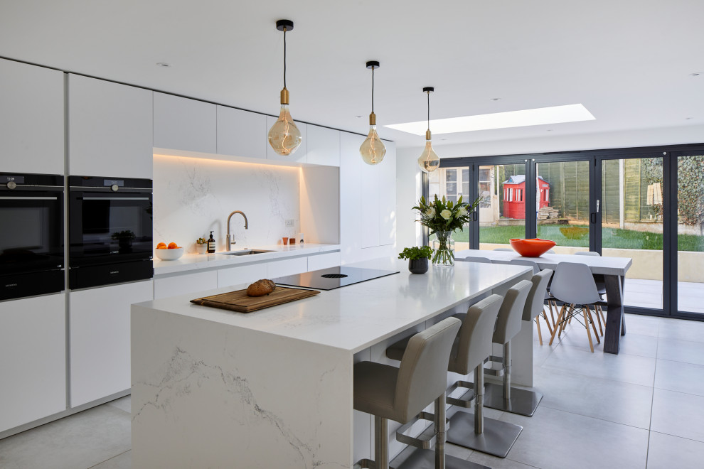 Large contemporary single-wall open plan kitchen in Kent with a submerged sink, flat-panel cabinets, white cabinets, engineered stone countertops, white splashback, engineered quartz splashback, stainless steel appliances, ceramic flooring, an island and white worktops.