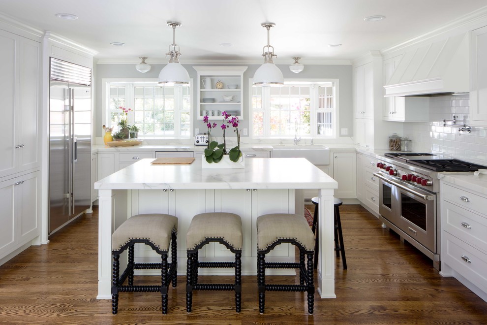 This is an example of a traditional u-shaped kitchen in San Francisco with a farmhouse sink, shaker cabinets, white cabinets, white splashback, subway tile splashback, stainless steel appliances, dark hardwood floors, with island, brown floor and marble benchtops.