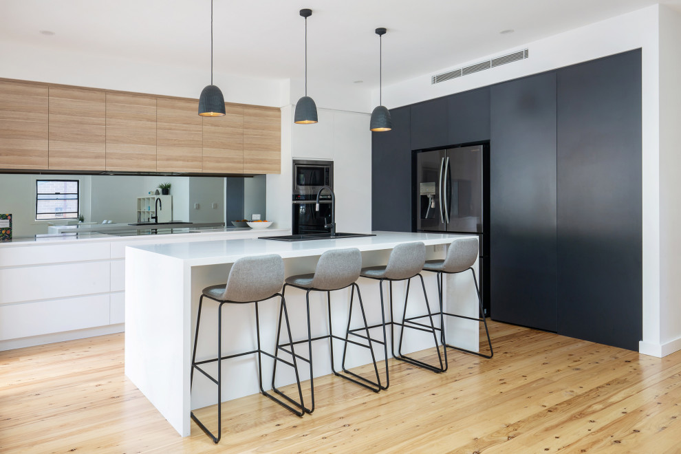 Mid-sized contemporary l-shaped open plan kitchen in Sydney with a drop-in sink, flat-panel cabinets, quartz benchtops, metallic splashback, mirror splashback, light hardwood floors, with island, white benchtop, black appliances and beige floor.