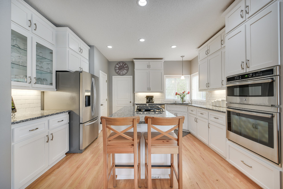 Mid-sized transitional l-shaped light wood floor and brown floor enclosed kitchen photo in Portland with a drop-in sink, shaker cabinets, white cabinets, quartz countertops, white backsplash, subway tile backsplash, an island and stainless steel appliances
