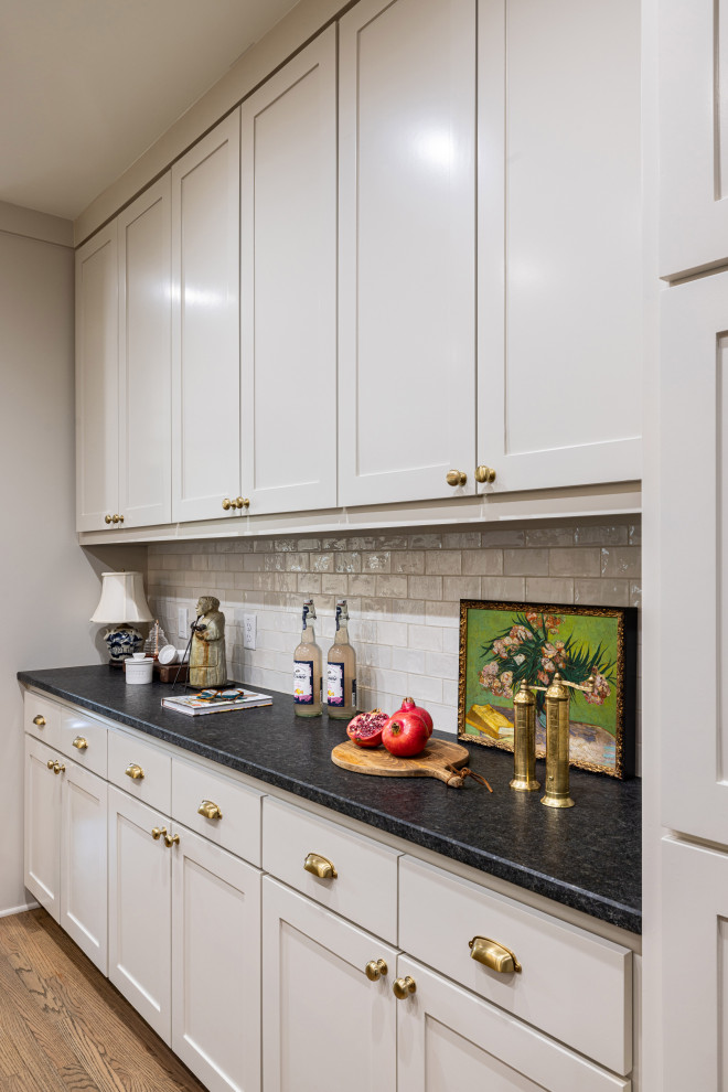 This is an example of a mid-sized transitional galley home bar in Dallas with no sink, shaker cabinets, beige cabinets, granite benchtops, beige splashback, subway tile splashback, medium hardwood floors, brown floor and black benchtop.