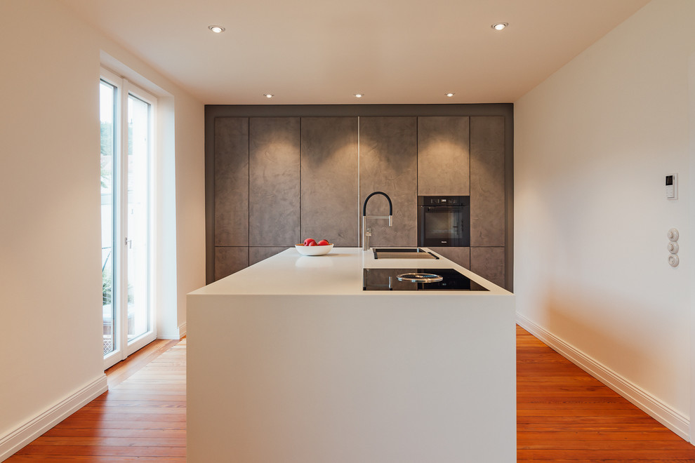 Design ideas for a mid-sized modern single-wall open plan kitchen in Other with an integrated sink, flat-panel cabinets, solid surface benchtops, grey splashback, black appliances, painted wood floors, with island, brown floor and white benchtop.