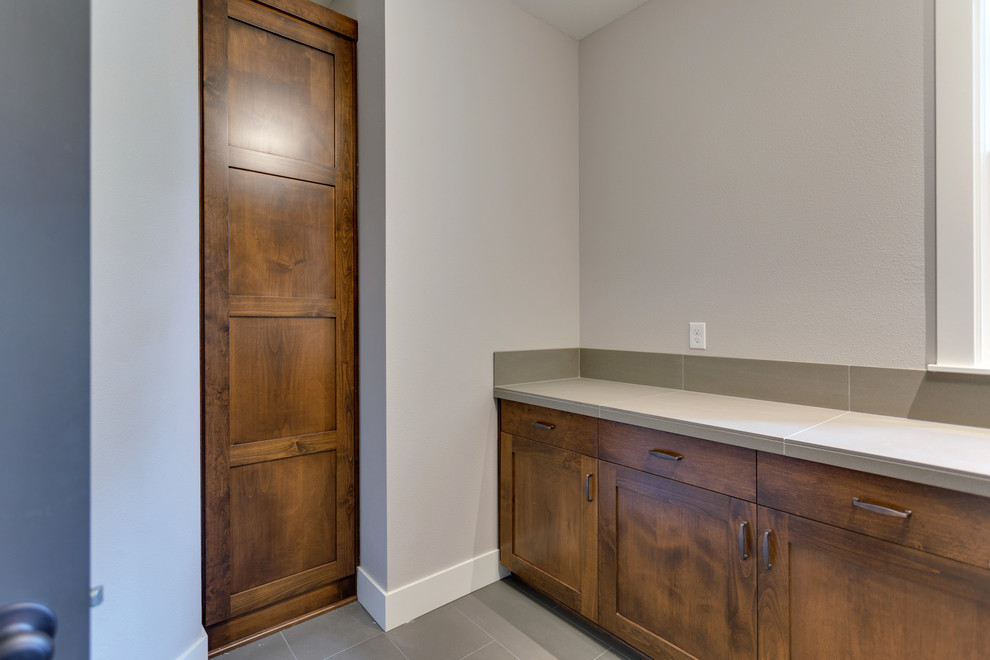 Mid-sized country l-shaped dedicated laundry room in Salt Lake City with an undermount sink, recessed-panel cabinets, dark wood cabinets, tile benchtops, grey walls, porcelain floors, a side-by-side washer and dryer, grey floor and grey benchtop.