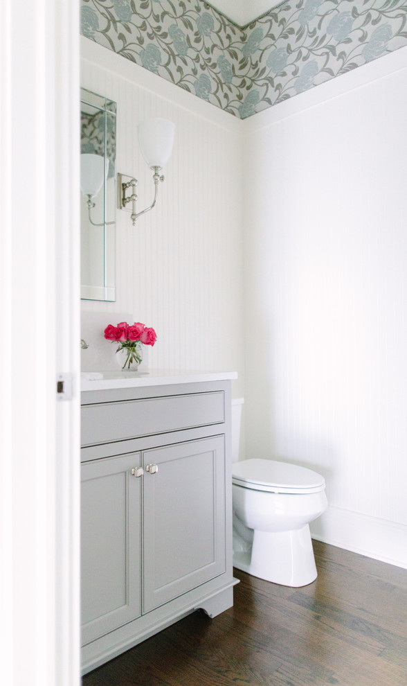 Photo of a mid-sized traditional powder room in Chicago with recessed-panel cabinets, grey cabinets, a two-piece toilet, multi-coloured walls, dark hardwood floors, engineered quartz benchtops and white benchtops.