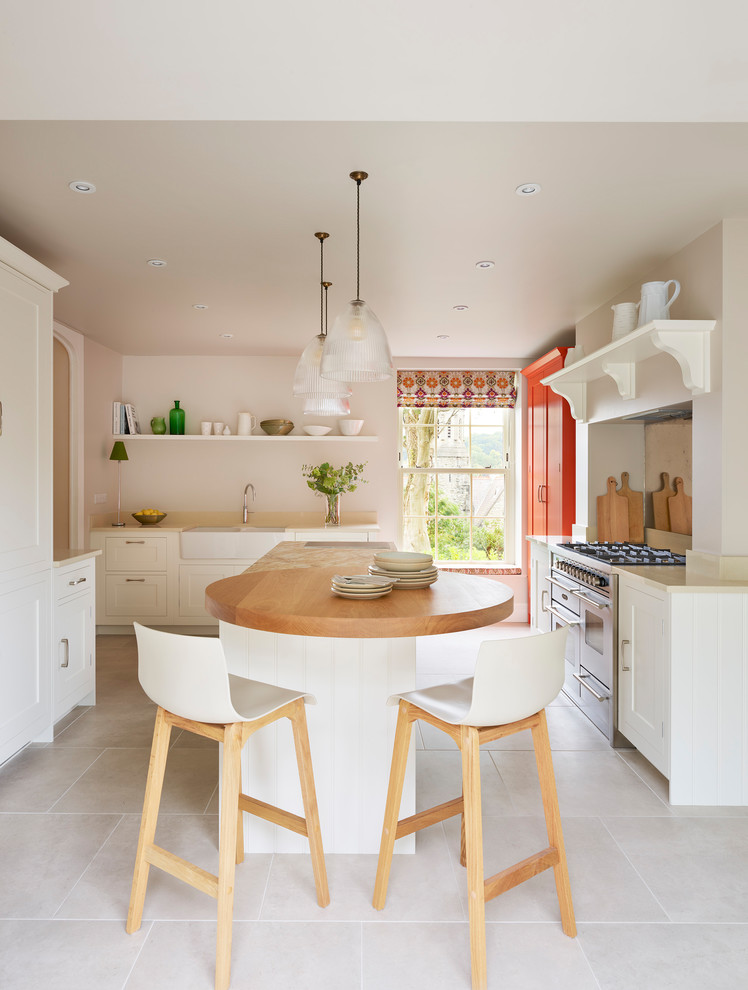 This is an example of a transitional u-shaped separate kitchen in London with a farmhouse sink, shaker cabinets, white cabinets, wood benchtops, stainless steel appliances, with island, beige floor and brown benchtop.