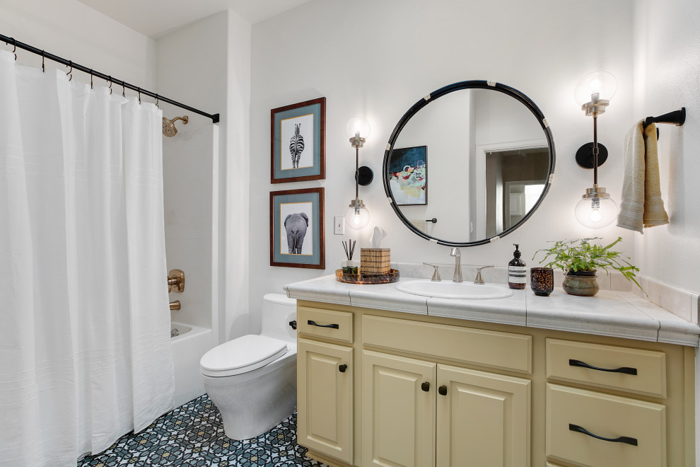 Photo of a mid-sized eclectic 3/4 bathroom in Portland with raised-panel cabinets, yellow cabinets, an alcove tub, a shower/bathtub combo, a one-piece toilet, multi-coloured tile, ceramic tile, white walls, porcelain floors, a drop-in sink, tile benchtops, multi-coloured floor, a shower curtain, beige benchtops, a single vanity, a built-in vanity and vaulted.
