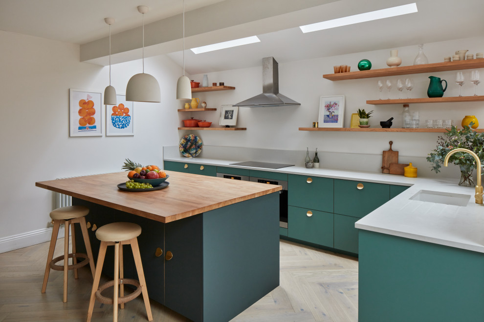 This is an example of a large eclectic l-shaped open plan kitchen in London with an integrated sink, flat-panel cabinets, green cabinets, quartz benchtops, grey splashback, engineered quartz splashback, black appliances, light hardwood floors, a peninsula and grey benchtop.