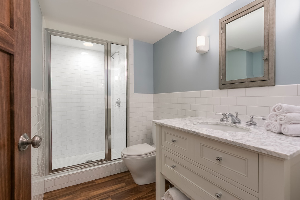 Photo of a mid-sized transitional 3/4 bathroom in Minneapolis with furniture-like cabinets, beige cabinets, an alcove shower, a one-piece toilet, white tile, subway tile, blue walls, medium hardwood floors, an undermount sink, marble benchtops, brown floor, a hinged shower door and multi-coloured benchtops.