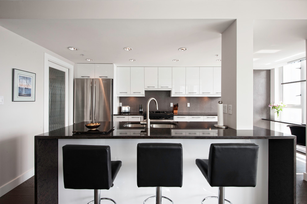 Photo of a contemporary galley kitchen in Vancouver with a double-bowl sink, flat-panel cabinets, white cabinets, quartz benchtops, grey splashback, stainless steel appliances, medium hardwood floors, with island and black benchtop.