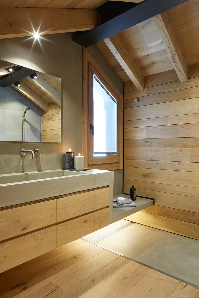Photo of a mid-sized country master bathroom in Other with a curbless shower, a two-piece toilet, light hardwood floors, flat-panel cabinets, light wood cabinets, grey walls, an integrated sink, concrete benchtops, brown floor and an open shower.