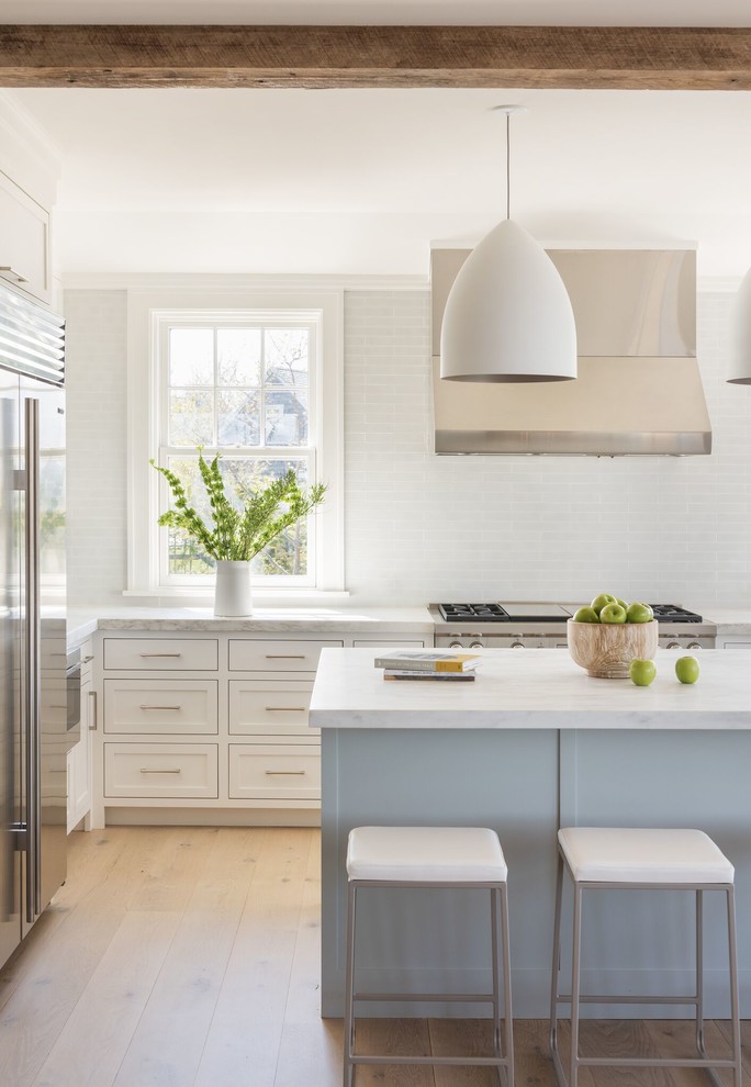 Inspiration for a mid-sized beach style u-shaped open plan kitchen in Providence with an undermount sink, beaded inset cabinets, white cabinets, marble benchtops, white splashback, glass tile splashback, stainless steel appliances, light hardwood floors, with island and beige floor.