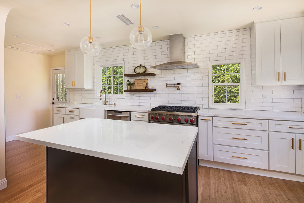 This is an example of a mid-sized country single-wall open plan kitchen in Los Angeles with a farmhouse sink, shaker cabinets, white cabinets, quartz benchtops, white splashback, porcelain splashback, stainless steel appliances, light hardwood floors, with island, brown floor and yellow benchtop.