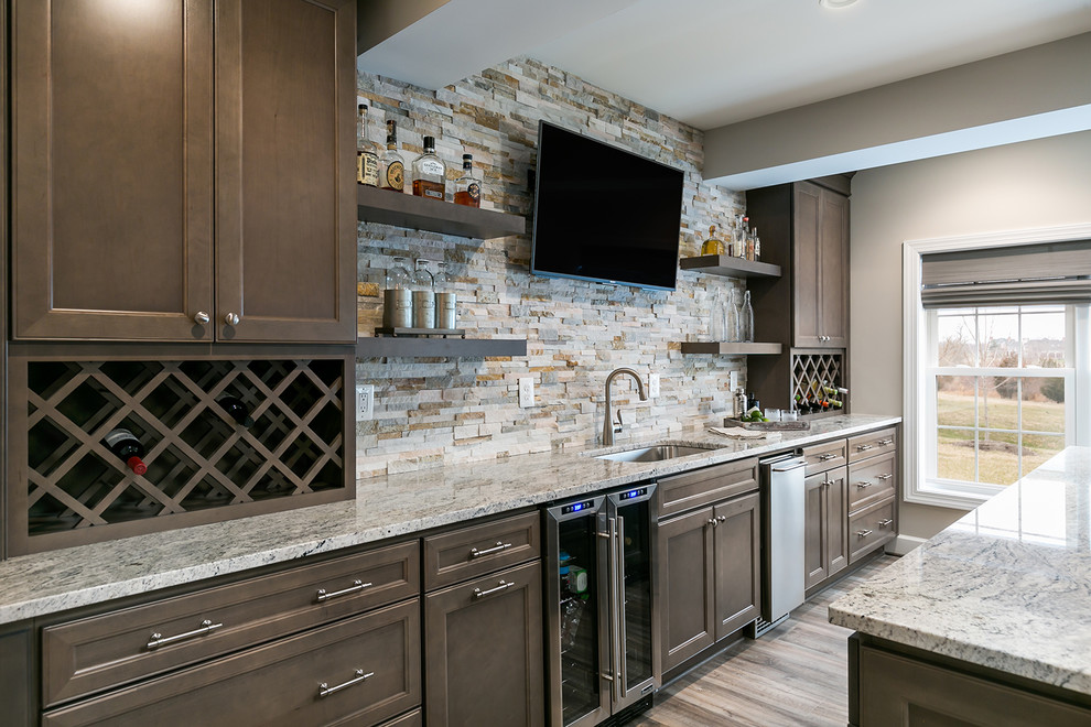 Photo of a large transitional single-wall seated home bar in DC Metro with recessed-panel cabinets, brown cabinets, granite benchtops, beige splashback, stone tile splashback, laminate floors, brown floor, an undermount sink and grey benchtop.