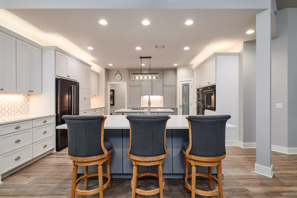 Example of a mid-sized transitional u-shaped vinyl floor and brown floor kitchen pantry design in Austin with a farmhouse sink, shaker cabinets, white cabinets, quartz countertops, white backsplash, marble backsplash, black appliances, two islands and white countertops