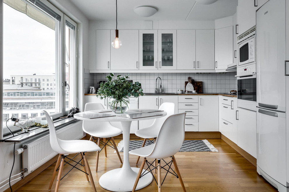 Mid-sized scandinavian l-shaped eat-in kitchen in Stockholm with flat-panel cabinets, white cabinets, wood benchtops, white splashback, white appliances, medium hardwood floors and no island.