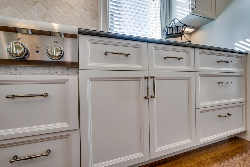 Mid-sized transitional l-shaped medium tone wood floor and brown floor open concept kitchen photo in Boston with a farmhouse sink, recessed-panel cabinets, white cabinets, soapstone countertops, beige backsplash, ceramic backsplash, stainless steel appliances and an island