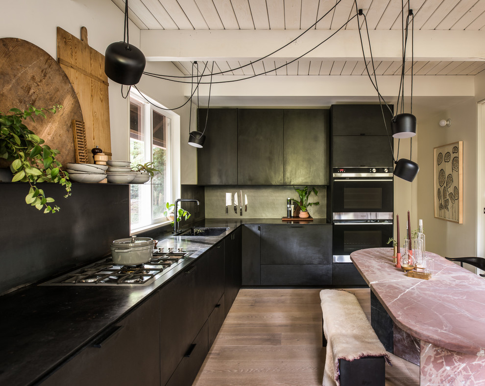 Contemporary l-shaped eat-in kitchen in Sacramento with flat-panel cabinets, black cabinets, black splashback, medium hardwood floors, brown floor and black benchtop.