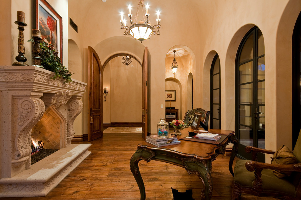 This is an example of an expansive mediterranean study room in Phoenix with beige walls, dark hardwood floors, a two-sided fireplace, a stone fireplace surround, a freestanding desk and brown floor.